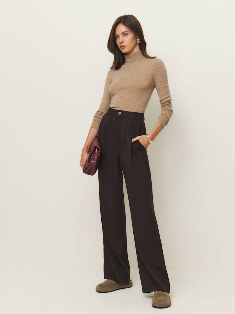 A woman wearing high-rise black wide-leg pants with a relaxed fit, paired with a beige turtleneck top, holding a maroon bag, and standing against a plain background.