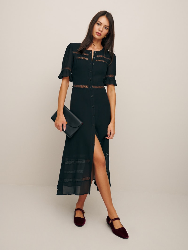 A woman wearing a black Petites Woodson Dress with lace accents, short sleeves, and a front slit, holding a black clutch and wearing burgundy shoes.