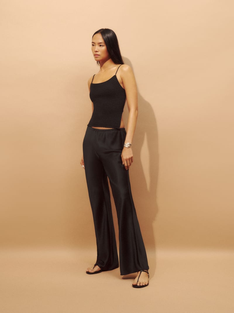 A woman wearing black satin Petites Gale Mid Rise Bias Pant with a black sleeveless top, standing against a beige background.