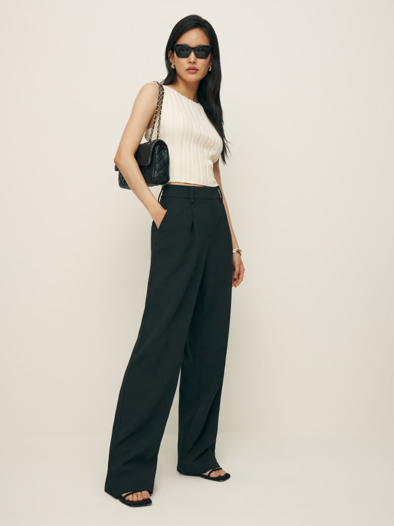 A woman wearing black high-waisted, pleated, wide-leg pants with a sleeveless cream top, black sunglasses, and a black quilted handbag.