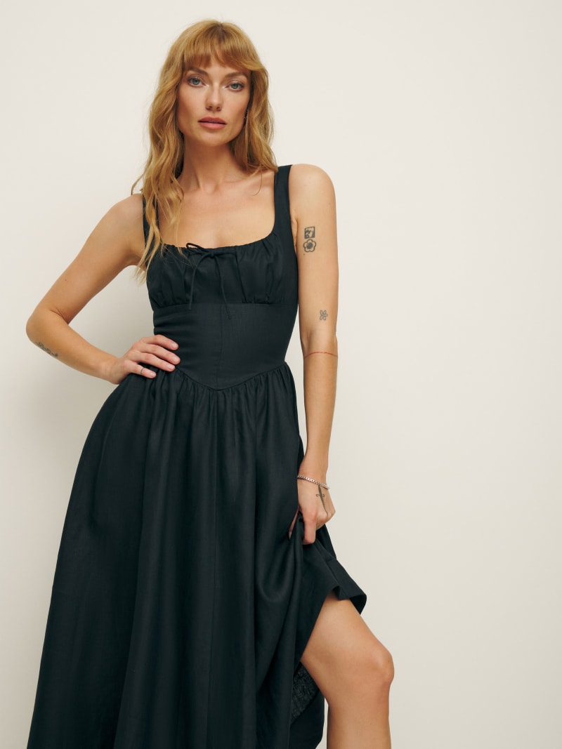 A woman wearing a black sleeveless linen maxi dress with a scoop neckline and front tie, standing against a plain background.