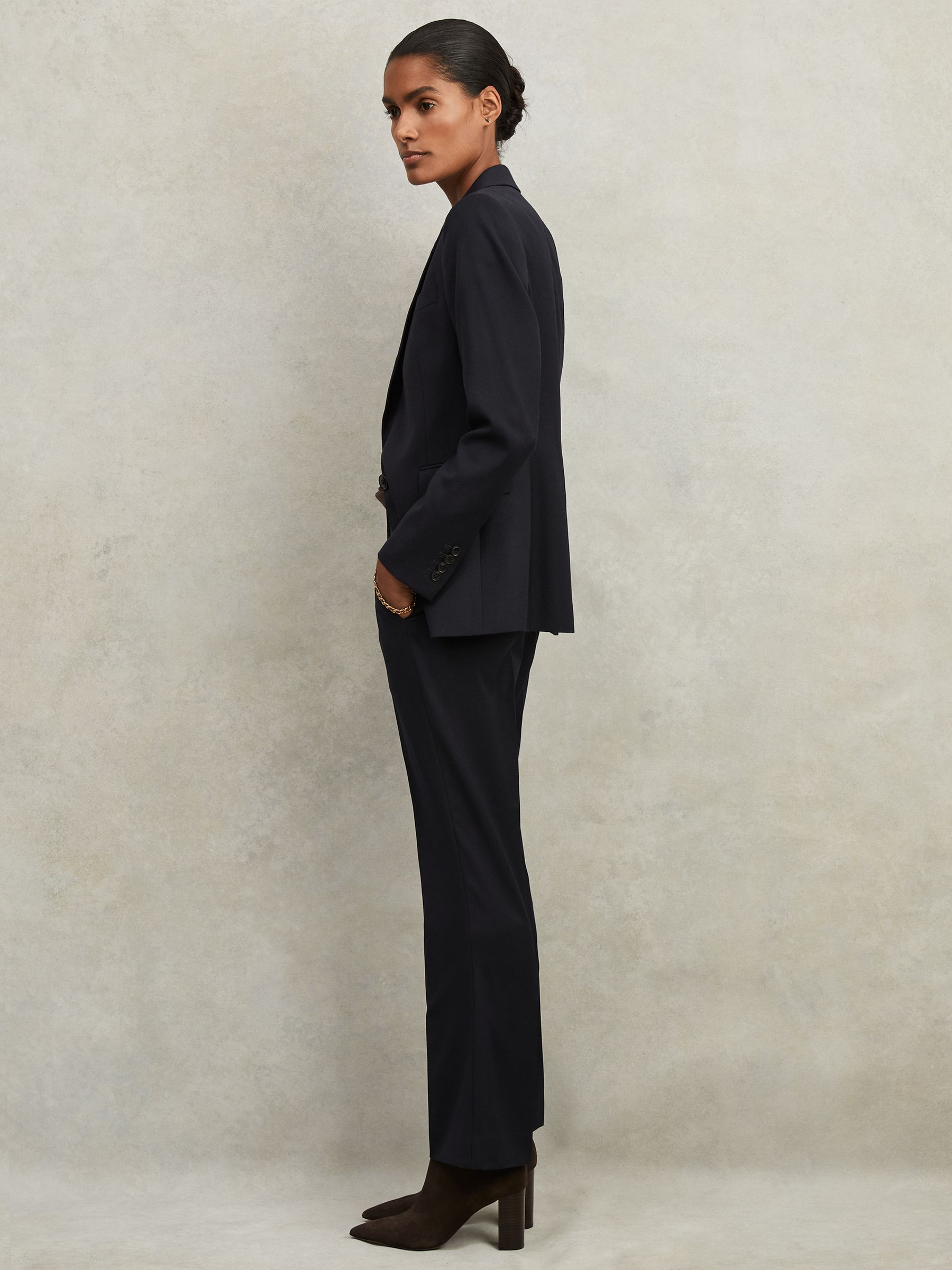 A woman wearing a navy suit blazer with notch lapels, single button closure, flap pockets, and buttoned cuffs, paired with matching trousers and brown high-heeled boots.