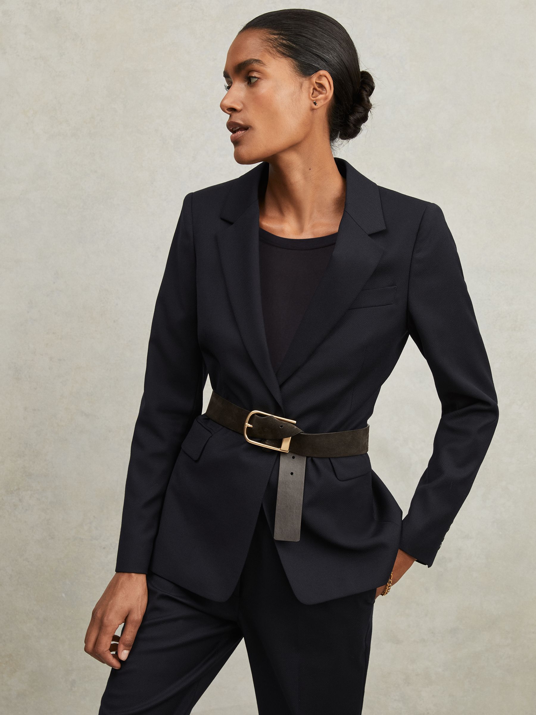 A woman wearing a navy single-breasted suit blazer with notch lapels, padded shoulders, and flap pockets.