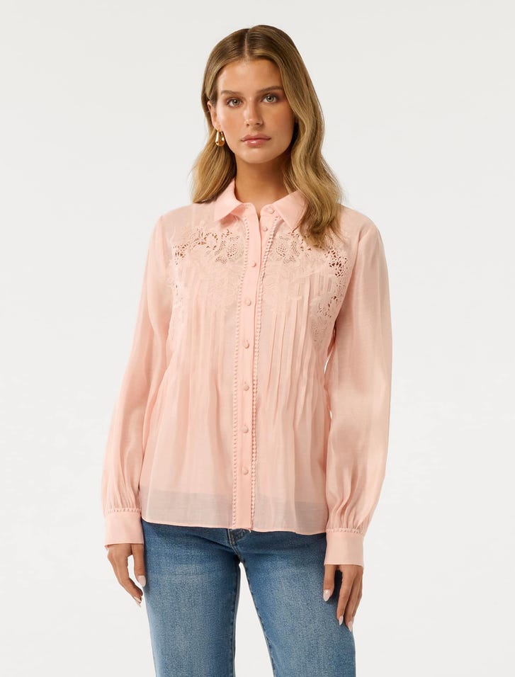 A woman wearing a light pink embroidered shirt with long sleeves, a collar, and button closure, featuring detailed embroidery on the upper chest area.