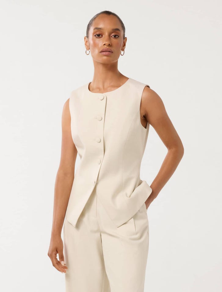 A woman wearing a sleeveless cream high neck waistcoat with a button-up front, paired with matching cream trousers.