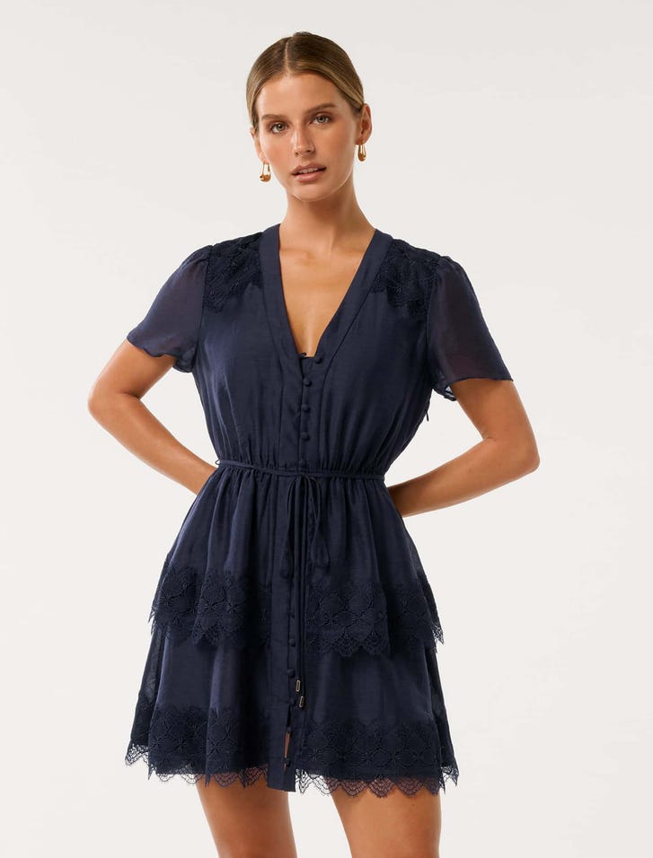 A woman wearing a navy blue mini dress with lace trim, short puffed sleeves, V-neckline, and a drawstring waist, with lace details on the shoulders and hem.