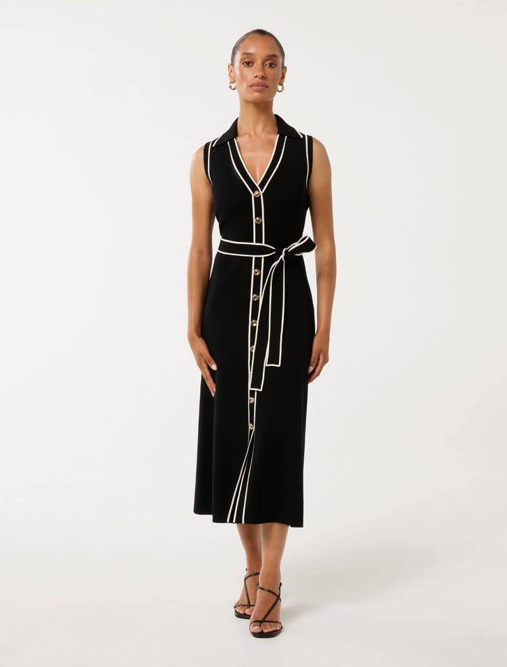 A woman wearing a sleeveless black midi dress with white piping, V-neckline, button-down front, and waist tie, paired with black strappy heels.