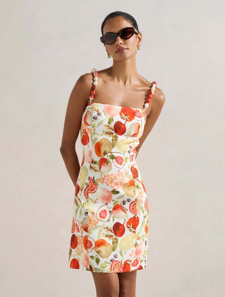 A woman wearing a sleeveless mini dress with a fruit print, beaded straps, and sunglasses.