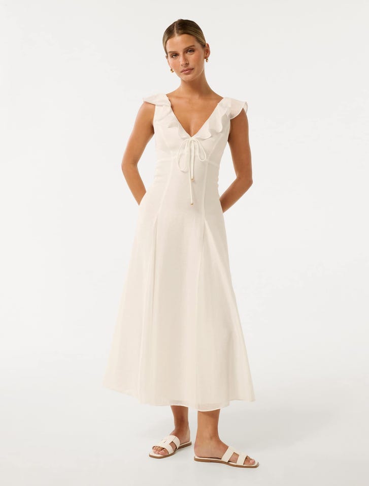A woman wearing a white midi dress with ruffled cap sleeves, a V-neckline with a tie, and a flared skirt, paired with white sandals.