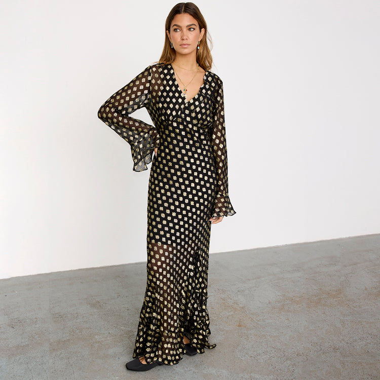 A woman wearing a black maxi dress with gold fleck print, sheer fabric, frill hem and cuffs, and a black slip underneath.