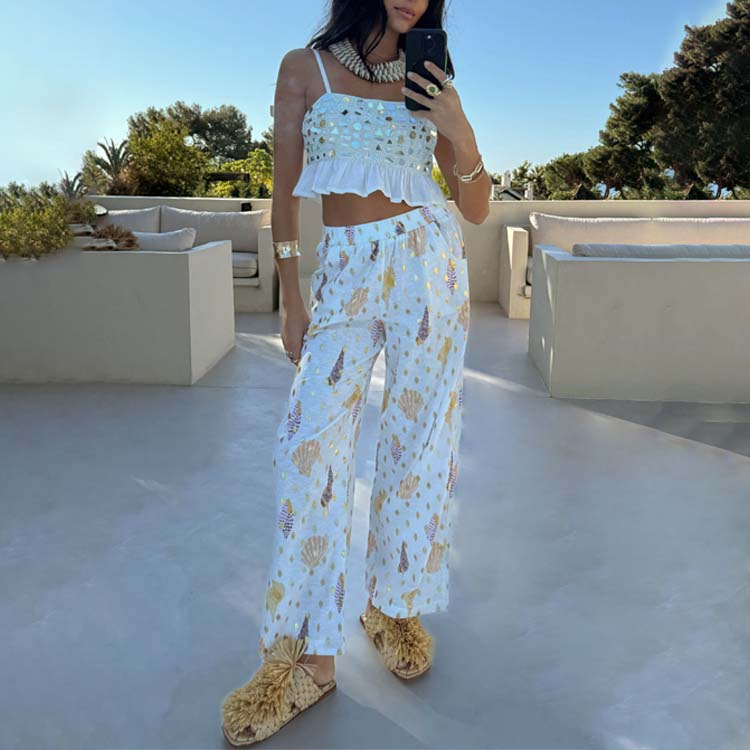 A woman wearing white Shells Elissa Trousers Petite with a shell print and gold flecks, paired with a white ruffled crop top, standing outdoors.