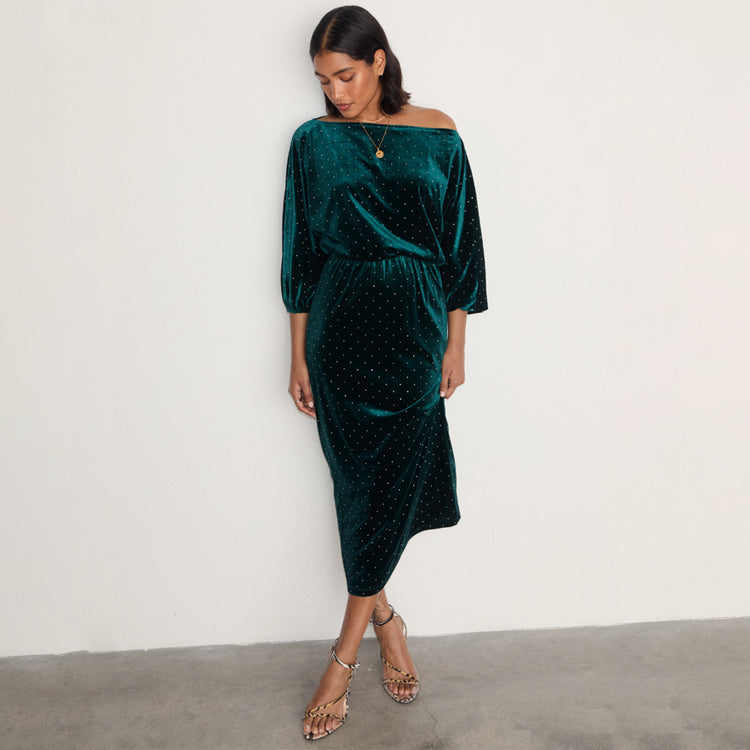 A woman wearing an emerald green velvet dress with a fitted skirt and a voluminous top, paired with high-heeled sandals.