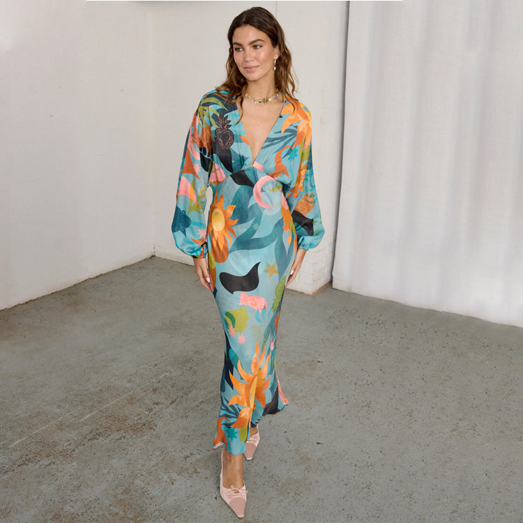 A woman wearing a multicoloured, long-sleeved maxi dress with a V-neckline, elasticated waist, and a vibrant print of various motifs.