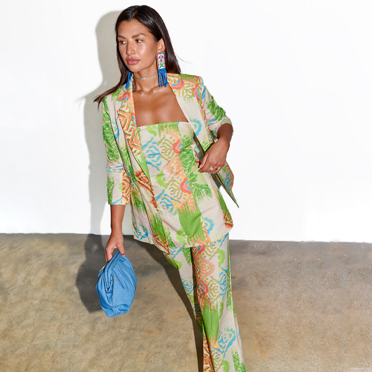 A woman wearing a colorful, patterned blazer with a matching top and pants, holding a blue clutch, standing against a white background.