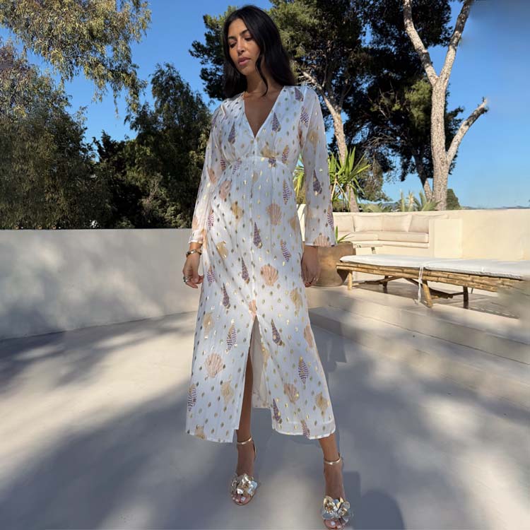 A woman wearing a white midi dress with a shell print and gold flecks, long sleeves, V-neckline, and a slit at the front, standing outdoors with trees and a blue sky in the background.