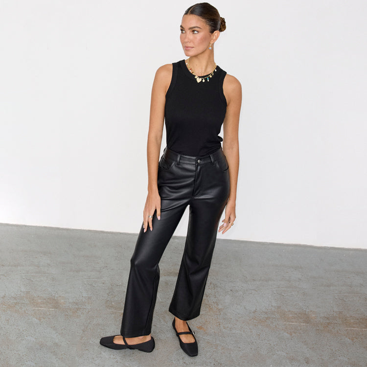A woman wearing black vegan leather trousers with a high waist and straight leg cut, paired with a black sleeveless top and black shoes.