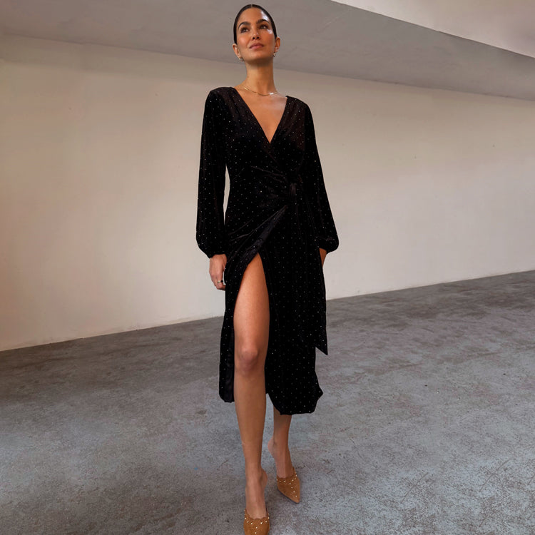 A woman wearing a black velvet midi dress with a deep V neckline, long sleeves, and a high slit on the side, standing in a minimalist indoor setting.