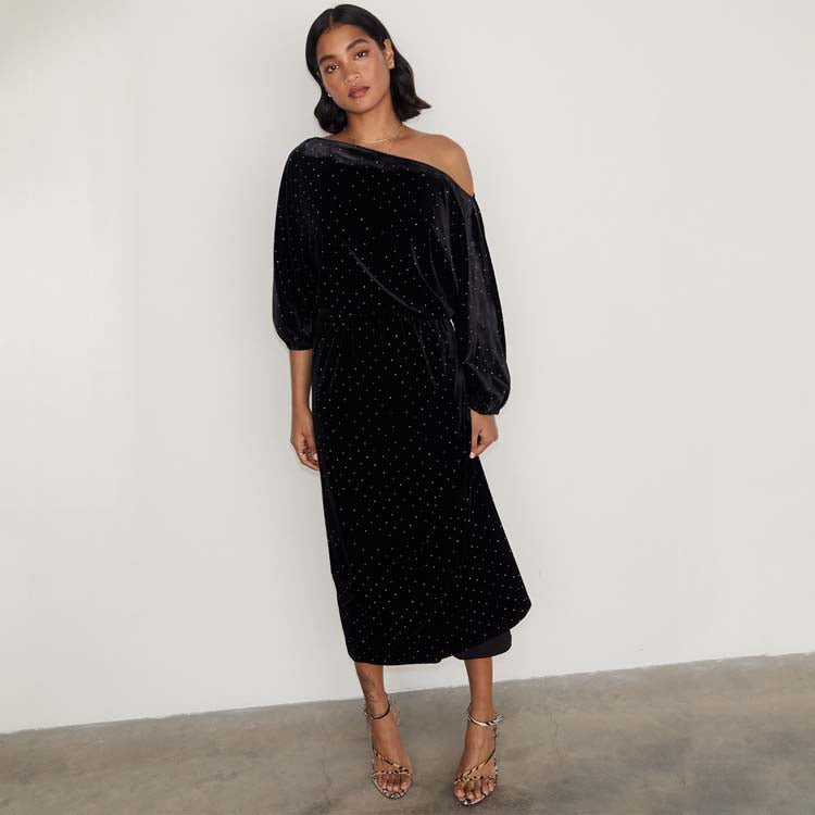 A woman wearing a black velvet off-the-shoulder dress with long sleeves and a dotted pattern, standing against a plain background.