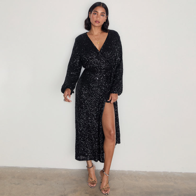 A woman wearing a black sequin wrap dress with a voluminous bodice, a high slit, and an adjustable waist, paired with high heels.