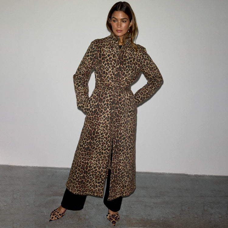 A woman wearing a long leopard print coat with a simple silhouette, front pockets, and contrast orange French seams, paired with black pants and leopard print shoes.