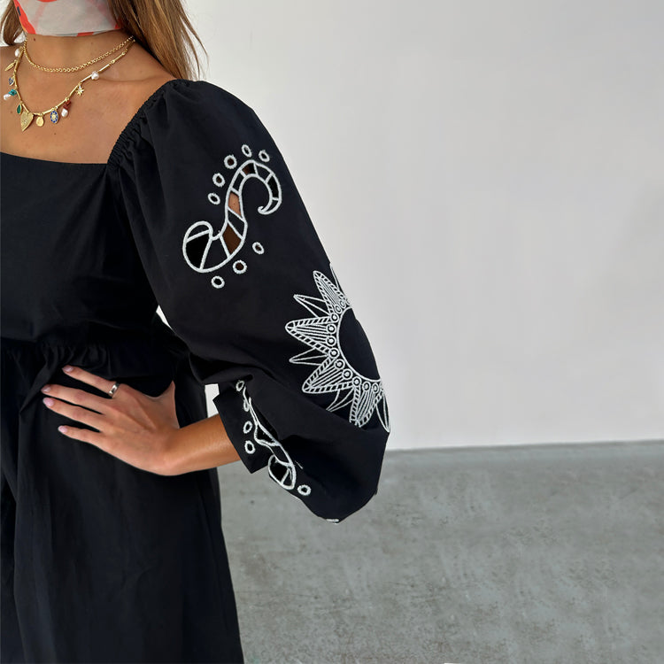 A black women's dress with embroidered cut-out motifs on the sleeves and a gathered neckline.