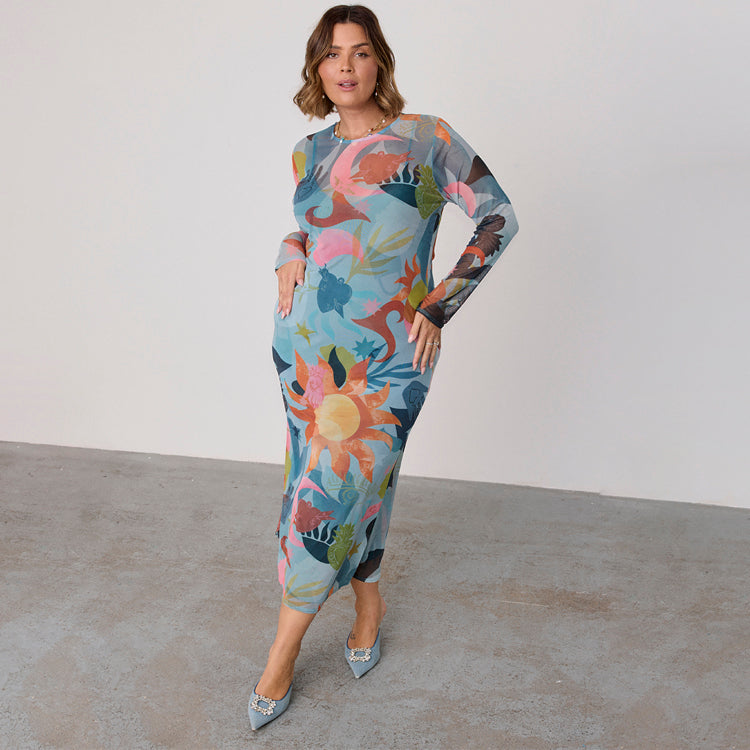 A woman wearing a multicoloured mesh dress with floral and motif print, long sleeves, and a fitted silhouette, paired with high heels.