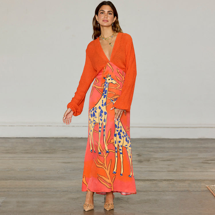 A woman wearing a bright orange dress with a giraffe print, crochet knit top, and a glossy viscose skirt, standing indoors.