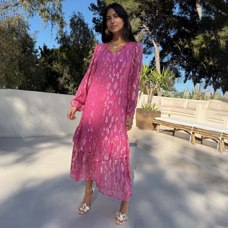 A woman wearing a pink dress with a flowy silhouette, pleated skirt, tassels on the neckline, and elasticated cuffs, standing outdoors with trees and a white wall in the background.