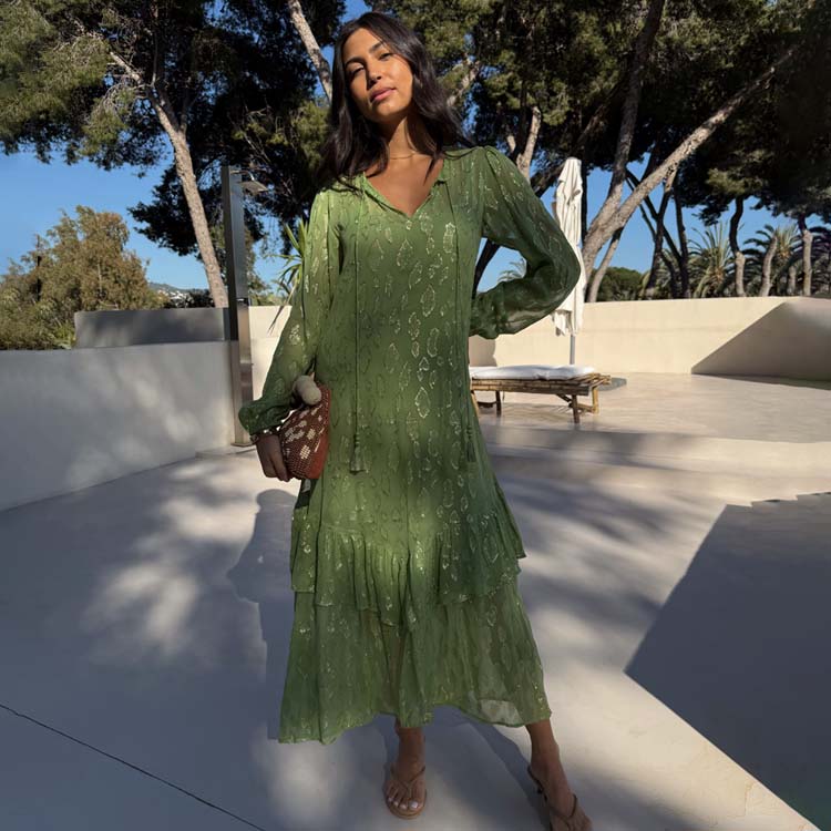 A woman wearing a flowy olive green dress with pleats, gold jacquard animal print, tassels on the neckline, and elasticated cuffs, standing outdoors.