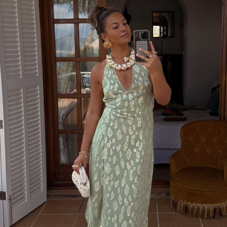 A woman wearing a sage green maxi dress with metallic gold jacquard pattern, halter neckline, and wrap tie fastening, accessorized with large pearl necklace and earrings.
