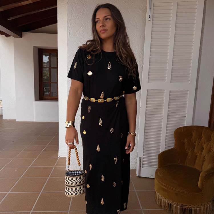 A woman wearing a black midi dress with gold shell and leaf embellishments, short sleeves, and a wrap tie skirt, accessorized with gold jewelry and a woven handbag.