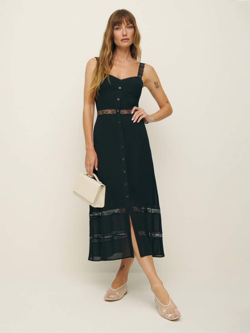 A woman wearing a black midi dress with lace accents, buttons down the front, and a slit at the bottom, holding a beige handbag and wearing transparent shoes.