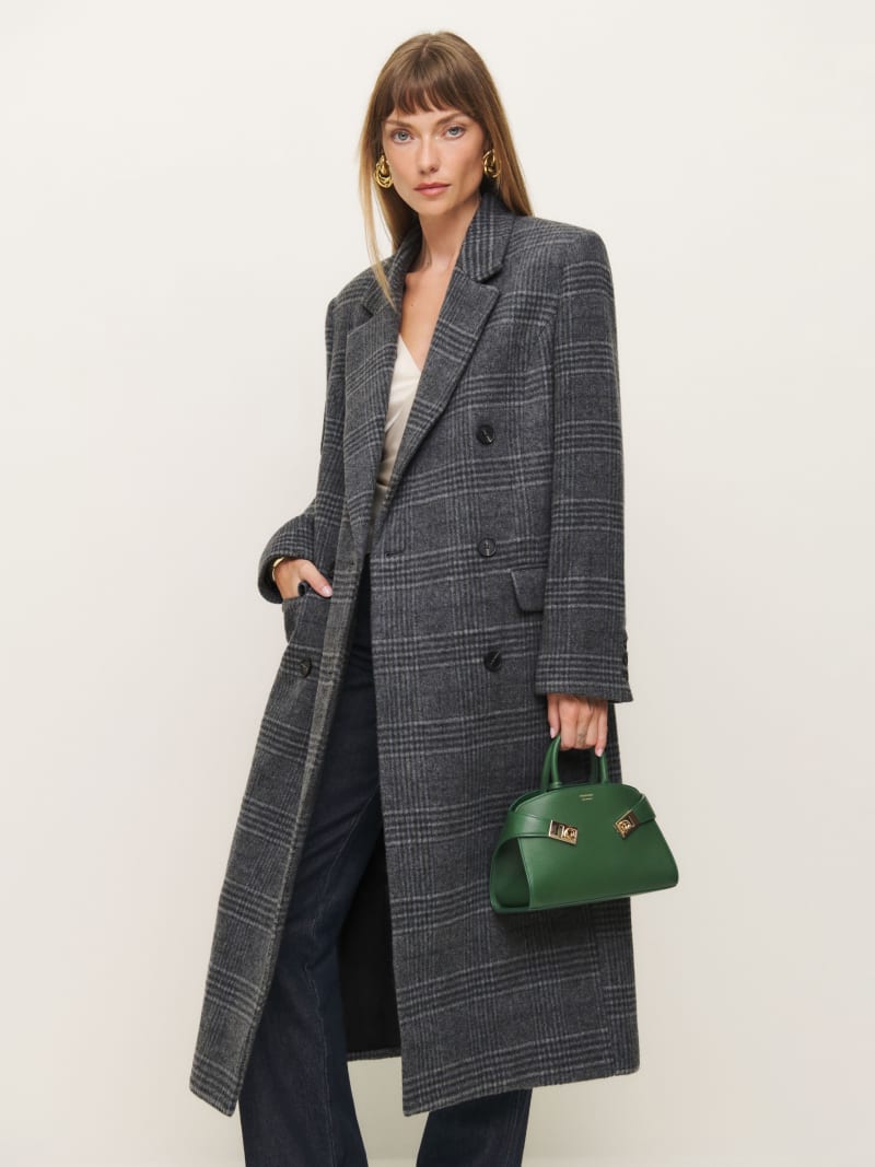 A woman wearing a long grey plaid coat with lapel collar and double-breasted buttons, holding a green handbag.