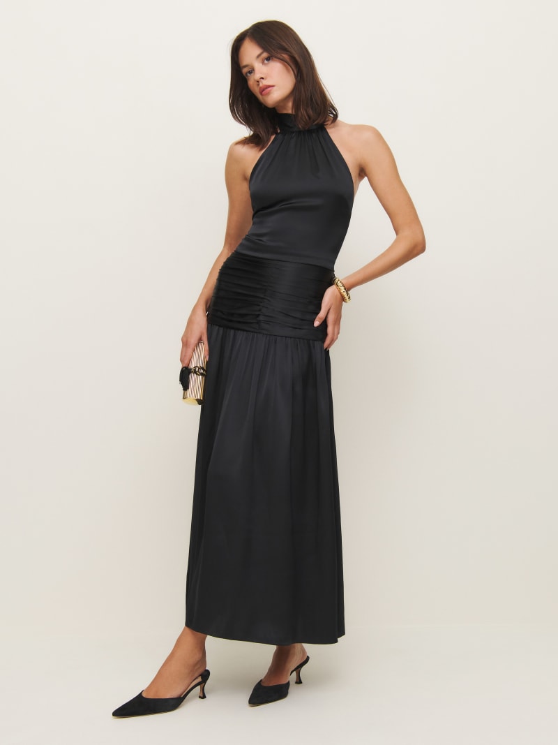 A woman wearing a black silk dress with long sleeves, a square neckline, and a smocked bodice, paired with black heels and holding a small clutch.