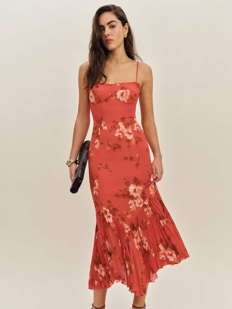 A woman wearing a coral pink floral midi dress with spaghetti straps, a sweetheart neckline with ruffles, and a pleated hem.