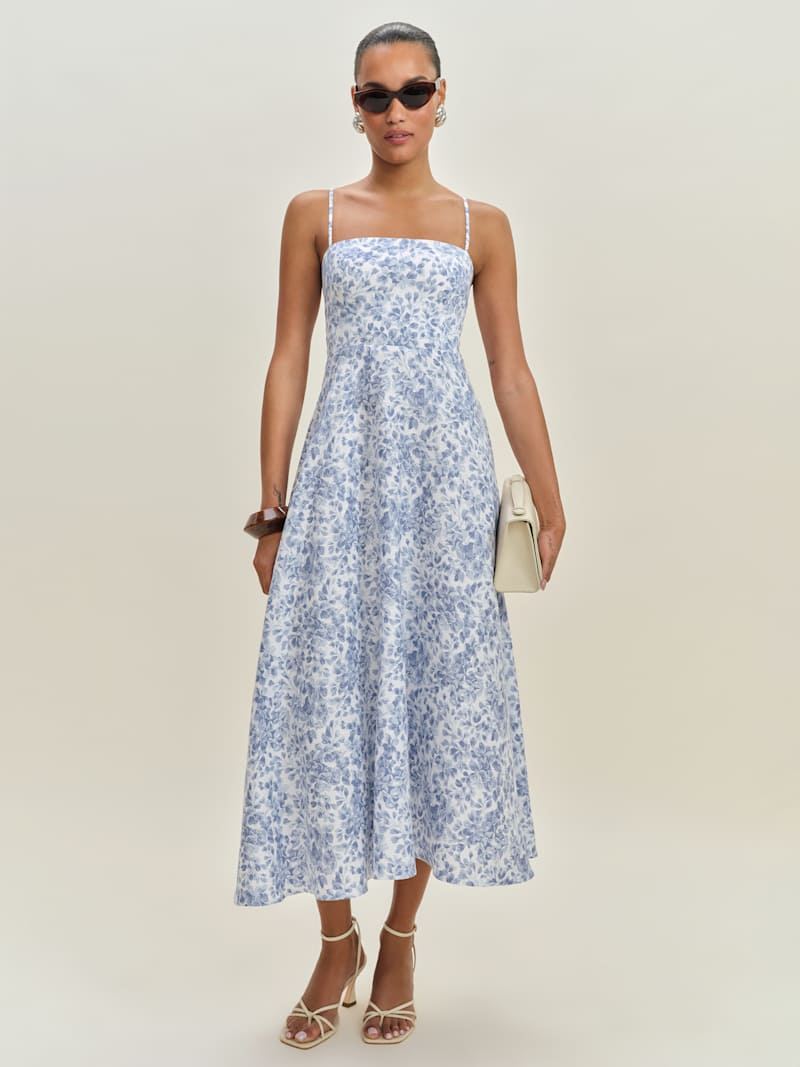 A woman wearing a sleeveless, blue and white floral linen dress with a square neckline, paired with beige high-heeled sandals, sunglasses, and a white clutch.