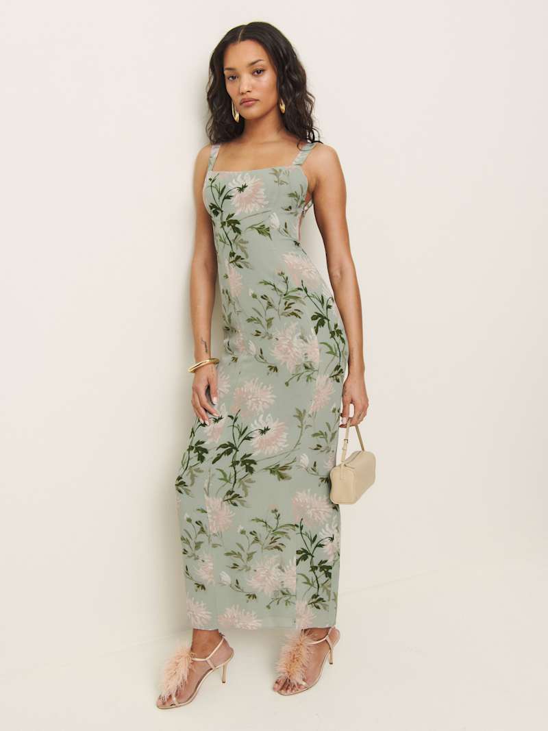 A woman wearing a sleeveless, full-length floral dress with a square neckline, open back with an adjustable tie, and a center back slit. She is holding a small beige handbag and wearing pink feathered heels.