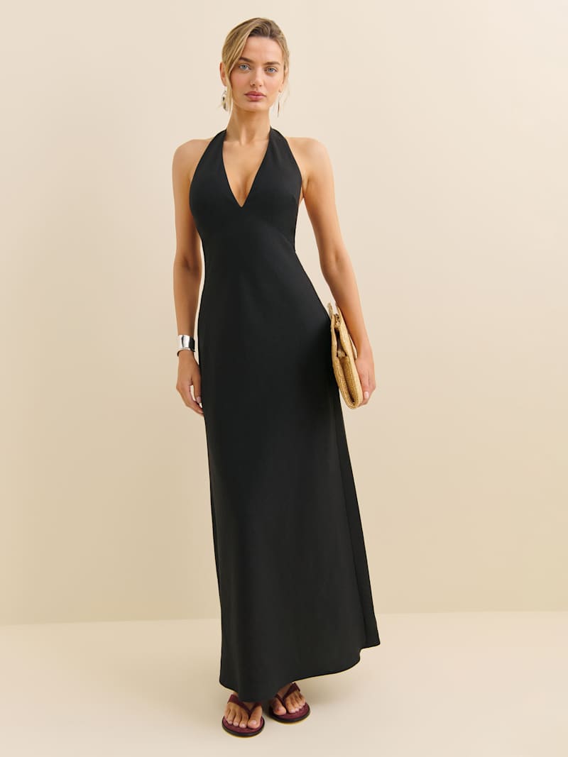 A woman wearing a black Petites Delilah Linen Dress with a halter neckline, maxi length, and sleeveless design, holding a beige clutch and wearing sandals.
