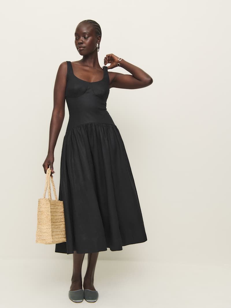 A woman wearing a black sleeveless linen dress with a drop waist and pleated skirt, holding a straw bag.