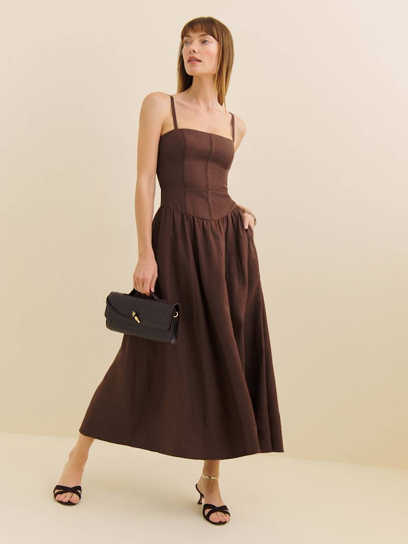 A woman wearing a brown sleeveless linen dress with a square neckline, drop waist, and pleated skirt, holding a black clutch and wearing black high-heeled sandals.