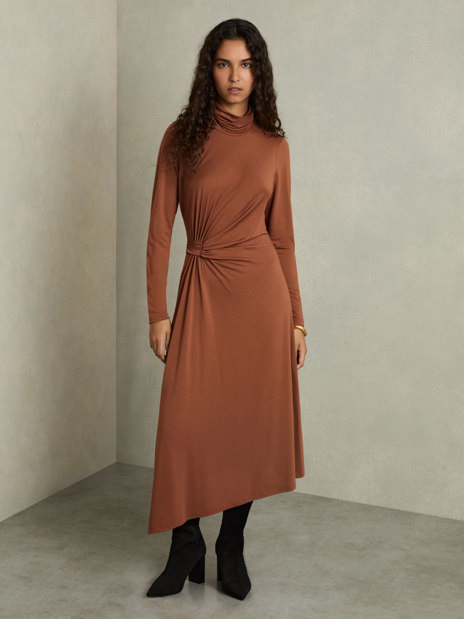 A woman wearing a camel-colored draped jersey midi dress with a roll neck and side slit.