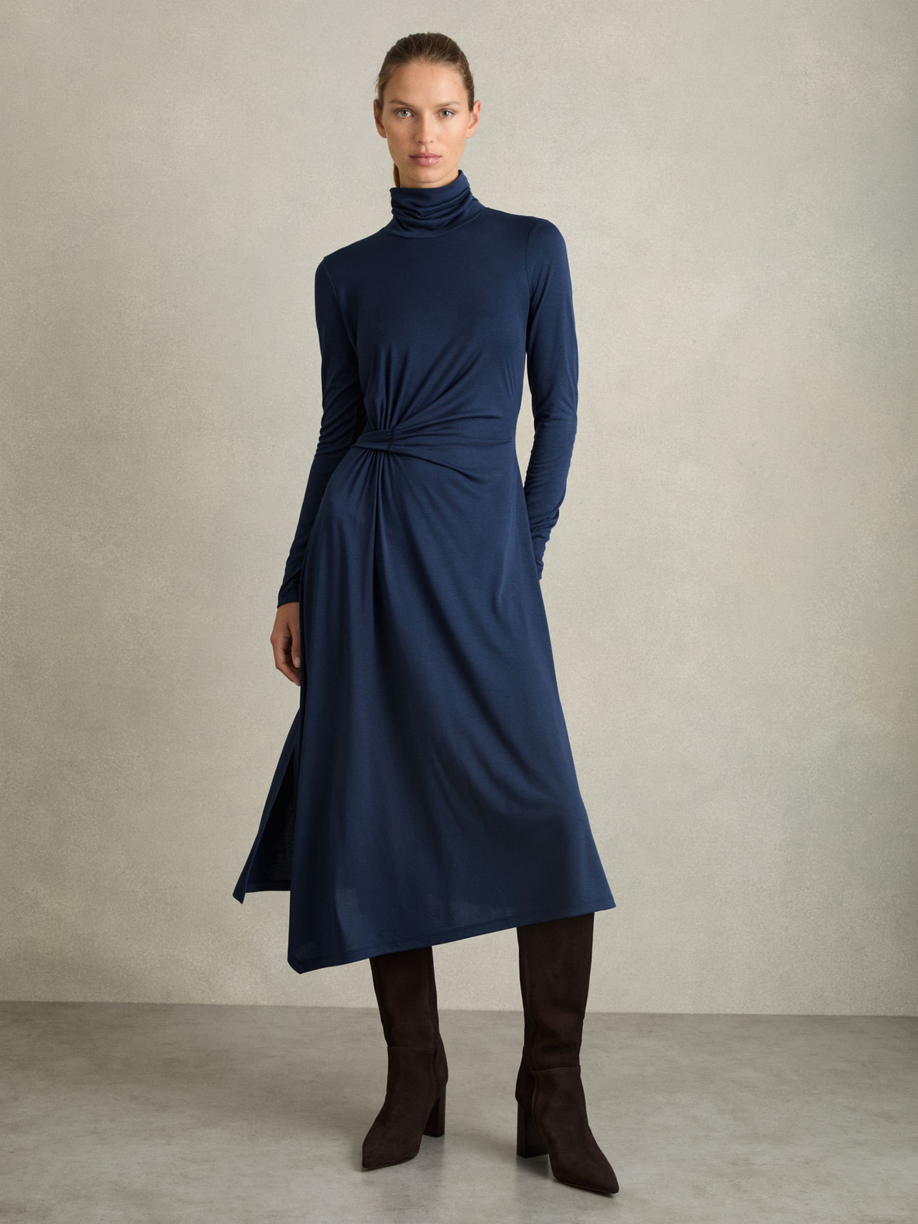 A woman wearing a blue draped jersey midi dress with asymmetric draping, a roll neck, and a side slit.