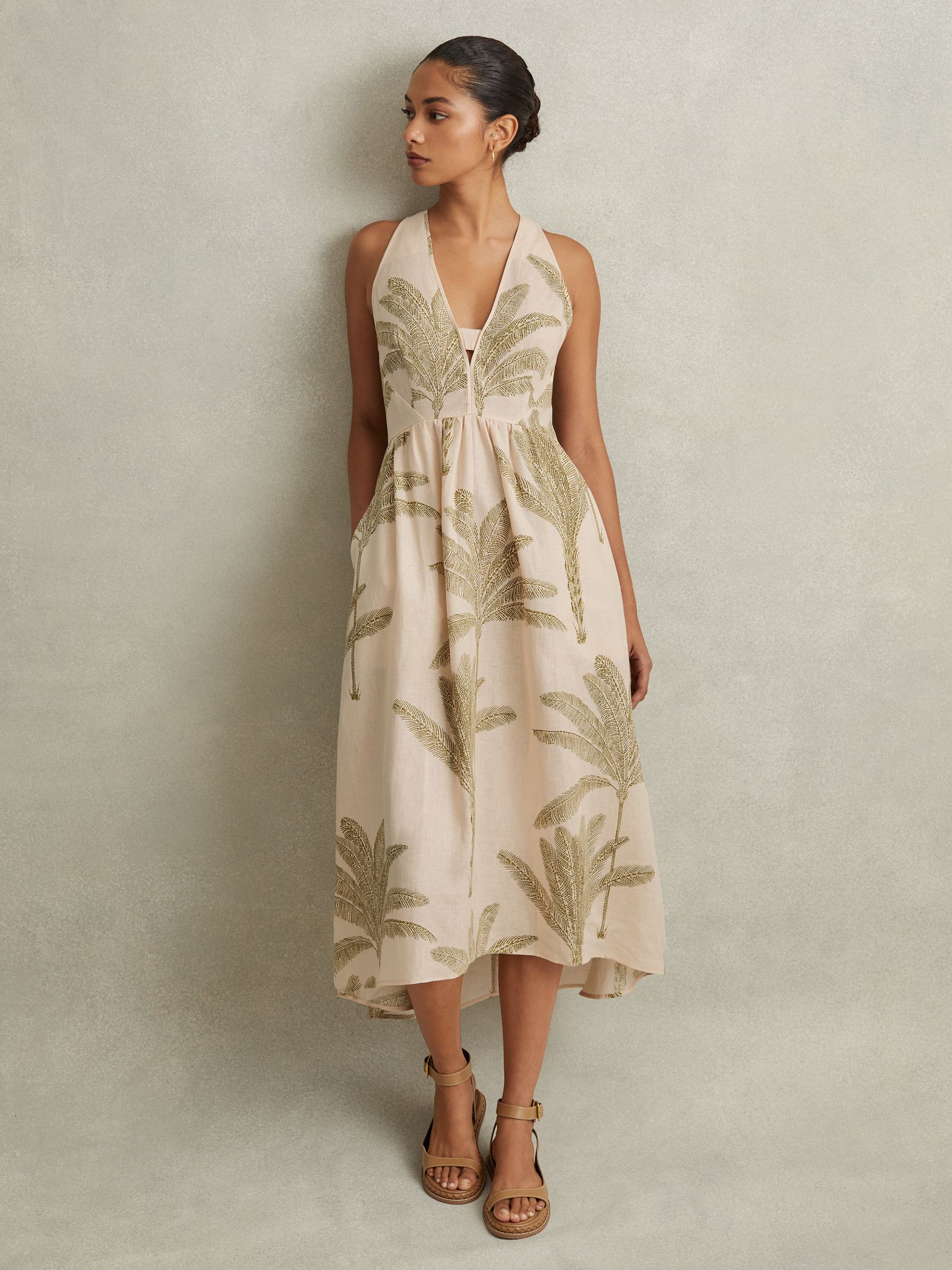 A woman wearing a sleeveless midi dress with a tropical print in neutral and green colors, featuring a V-neckline and side pockets.