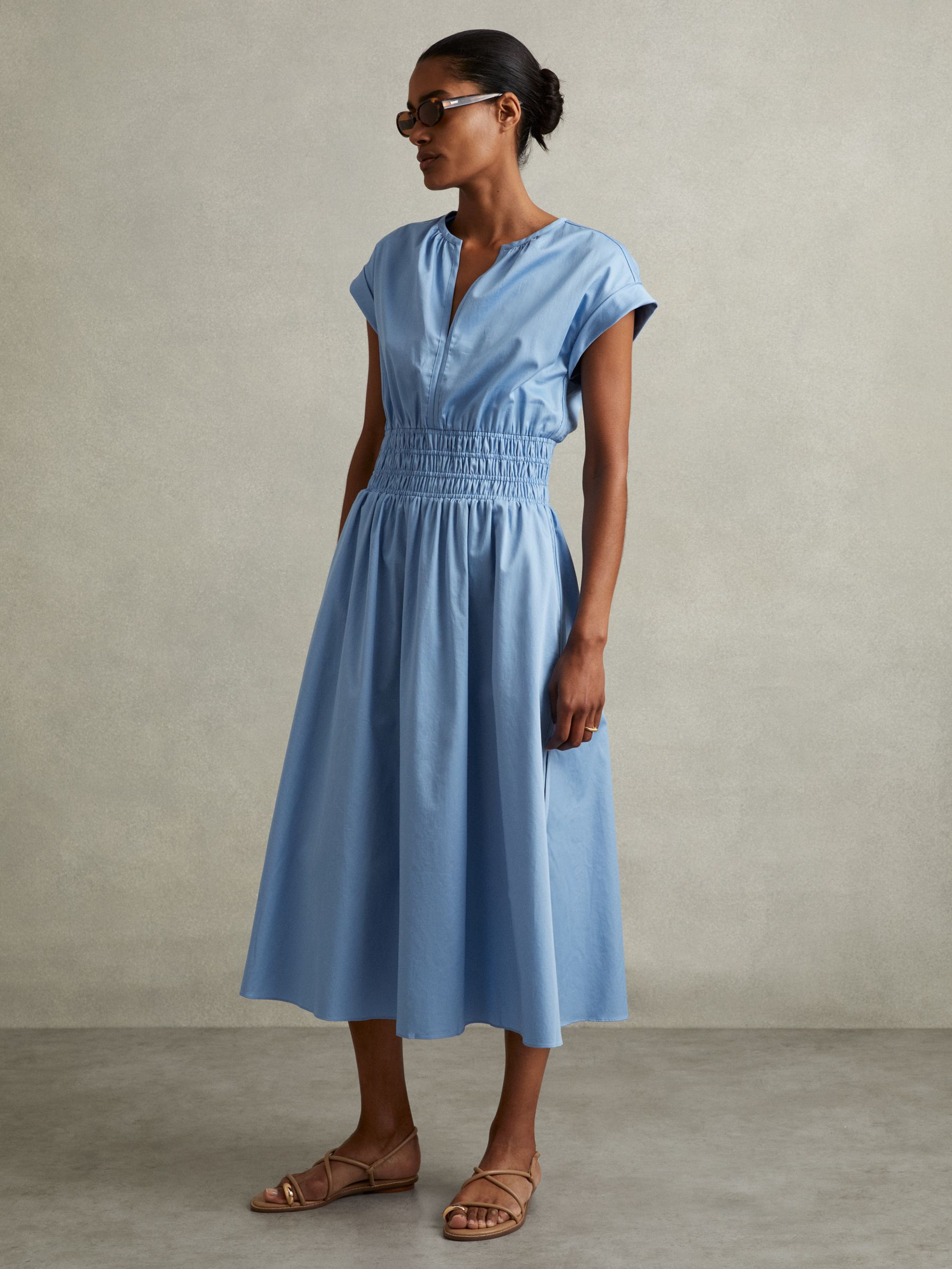 A woman wearing a blue cotton midi dress with ruched waist, V-neckline, rolled sleeves, and side splits.