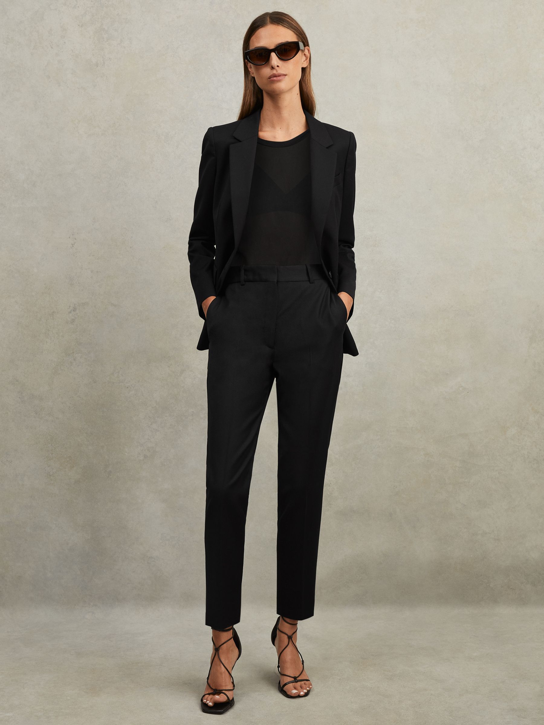 A woman wearing black tapered suit trousers with a blazer, black top, and high heels, standing with hands in pockets.