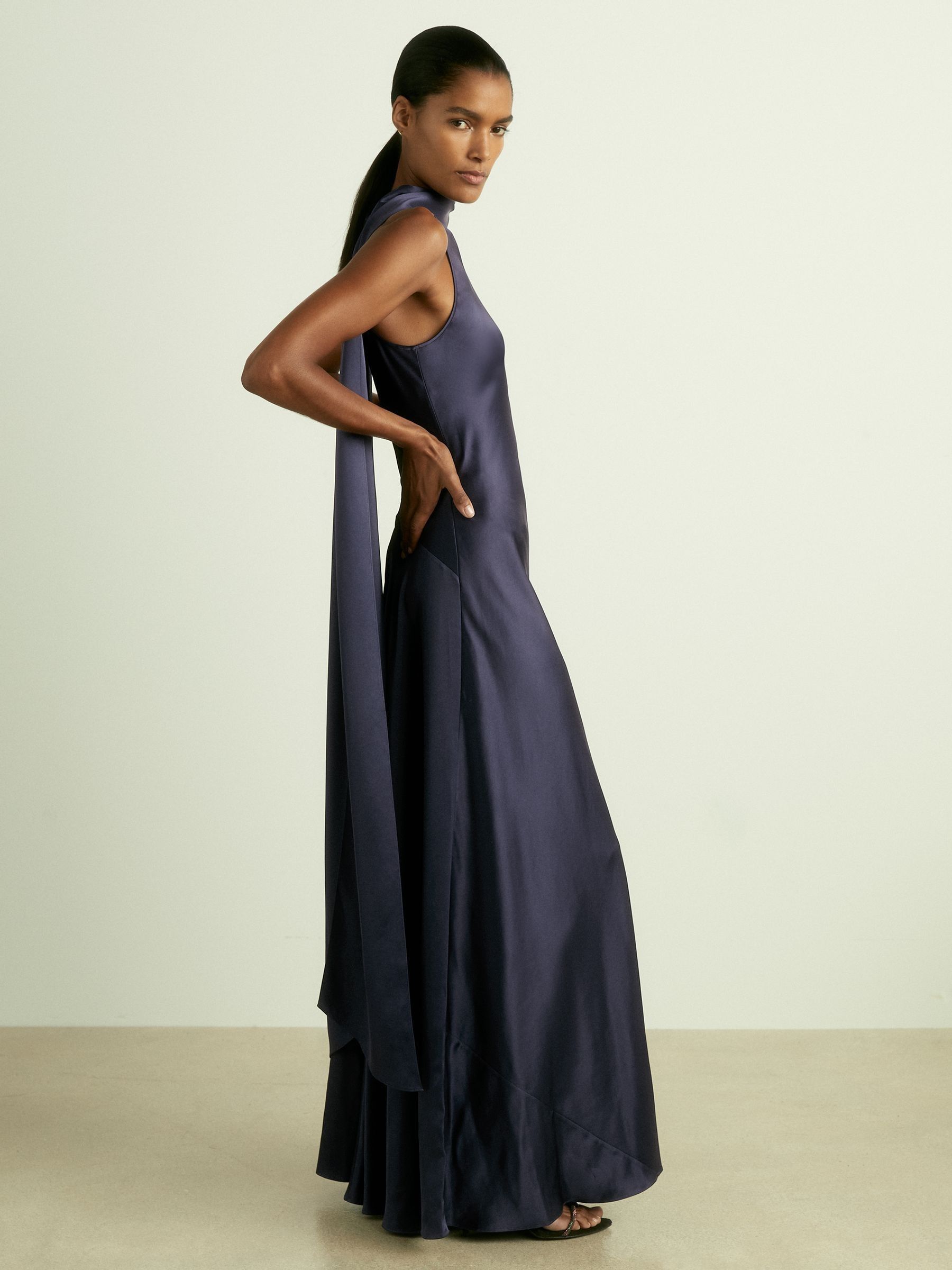 A woman wearing a long, indigo satin cape dress with a high neck and a matching scarf draping down the back, creating a split cape effect.