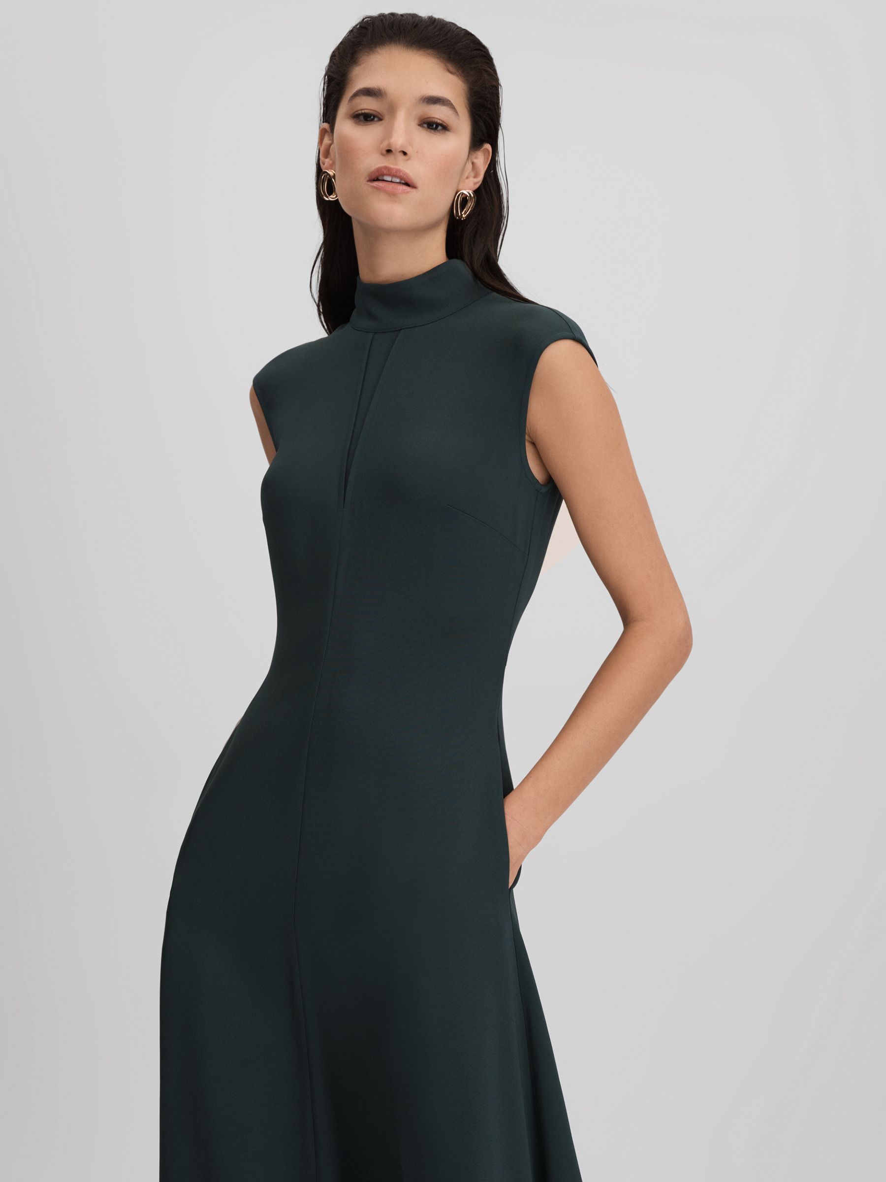 A woman wearing a dark green fitted asymmetric midi dress with a high neckline, notch detail, and back zip and tie fastening.