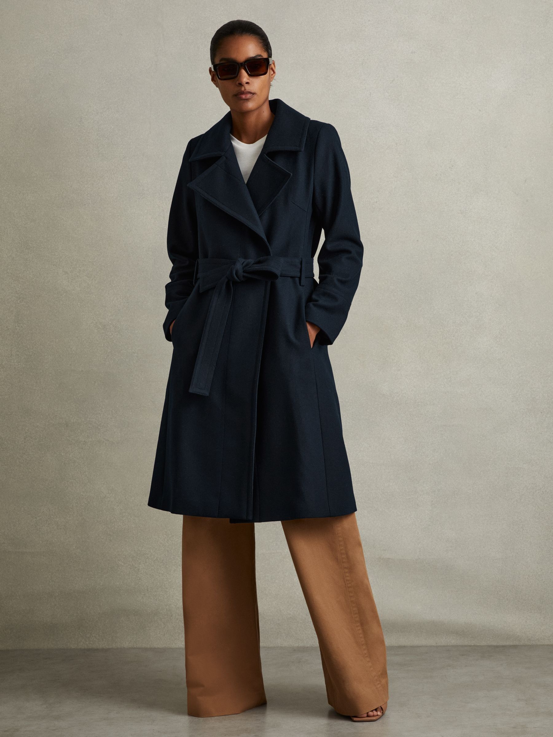 A woman wearing a teal wool blend belted double-breasted coat with a notched collar, knee-length hem, and satin lining. The coat has darts at the bust, inseam pockets, and concealed buttons.