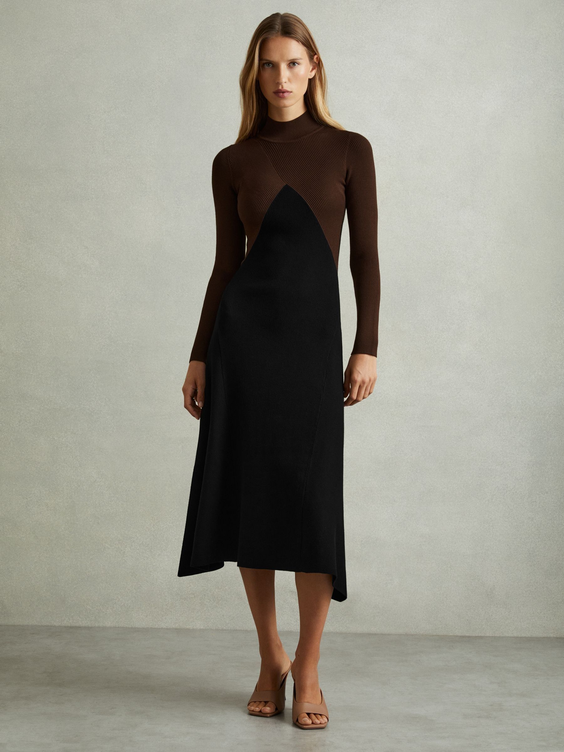 A woman wearing a colourblock ribbed midi dress with a high neckline, long sleeves, and dipped hem, featuring chocolate and black colour blocks.