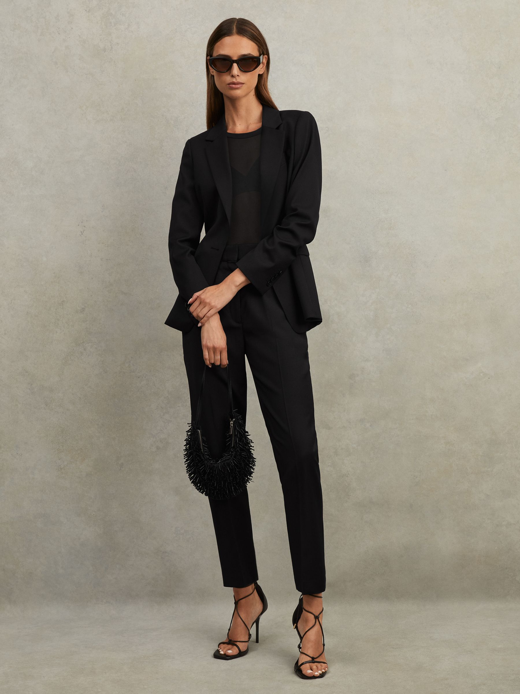 A woman wearing black tailored suit trousers with a mid-rise, pressed creases, side-slip pockets, and a slim, tapered leg, paired with a black blazer and high heels.
